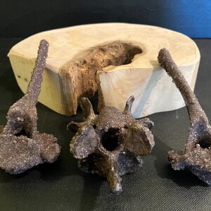 May include: Three dark purple amethyst crystal clusters, with a sparkling, textured surface, displayed on a wooden slab. The crystals are set against a dark background. The wooden slab has a natural edge.