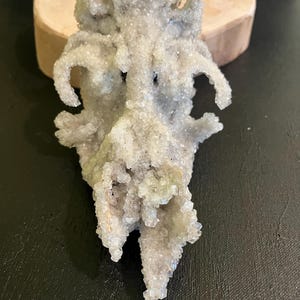 May include: A light grey, crystalline mineral specimen with a textured surface. The mineral has a unique, organic shape with multiple protrusions and a rough, bumpy texture. The specimen is displayed on a wooden base against a dark background.