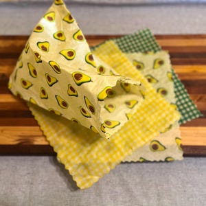 May include: A collection of reusable food wraps in various patterns. One wrap is folded into a triangular shape and features a yellow background with avocado illustrations. Other wraps have yellow gingham, avocado, and green gingham patterns.
