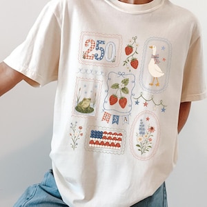 Puede incluir: Camiseta color crema con varios sellos ilustrados. Los diseños incluyen un sello "250", fresas, un ganso, una rana, la bandera estadounidense y elementos florales. La camiseta tiene un corte holgado.