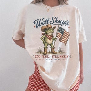 Könnte beinhalten: Cremefarbenes T-Shirt mit einer Cartoon-Froschfigur mit Cowboyhut, Stiefeln und rotem Halstuch, die eine amerikanische Flagge hält. Das Shirt trägt den Text "Well Sheeyit" über dem Frosch und "250 YEARS, STILL KICKIN' 1776 2026" darunter.