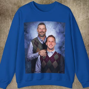 May include: A royal blue sweatshirt featuring a photograph of two people wearing patterned sweaters. The photo shows two people smiling and standing next to each other. The person on the left is wearing a green and brown patterned sweater and the person on the right is wearing a red and white patterned sweater.