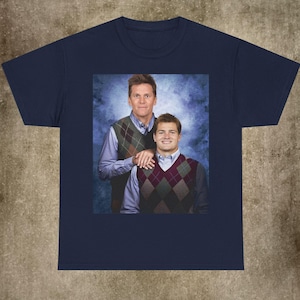 Peut inclure: Un t-shirt bleu marine avec une impression graphique centrée représentant deux personnes posant pour un portrait. Tous deux portent des chemises à col bleu clair sous des gilets en tricot argyle marron, vert et bordeaux sur fond bleu.