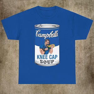 Op de afbeelding: Een koninklijk blauw t-shirt met een afbeelding van een Campbell's soepblik met een man die een kom soep eet. Het blik heeft de tekst "Knee Cap Soup" erop.