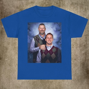 May include: Royal blue t-shirt with a framed image of two people wearing argyle vests. The image shows two men, one with a hand on the other's shoulder, against a blue and white background.