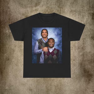May include: Black t-shirt featuring a portrait of two men wearing argyle vests over button-down shirts. The image has a blue and white background. The men are smiling and appear to be posing for a photo.
