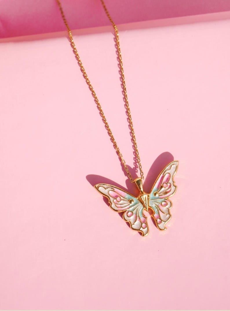 Pu&ograve; includere: Una collana con catena dorata con un ciondolo a forma di farfalla. La farfalla &egrave; bianca con accenti rosa e blu.