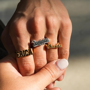 May include: Three gold rings with names engraved on them. The rings are worn on the fingers of a person's hand. The names on the rings are "Erica", "Chanda", and "Dominica".