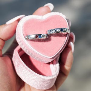 Peut inclure: Deux bagues en argent avec de petites pierres précieuses serties au centre. Les bagues sont dans un écrin en velours rose en forme de cœur.