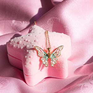 Pu&ograve; includere: Una collana con ciondolo a forma di farfalla dorata con dettagli in smalto blu e bianco, riposante in una scatola a forma di stella in velluto rosa.