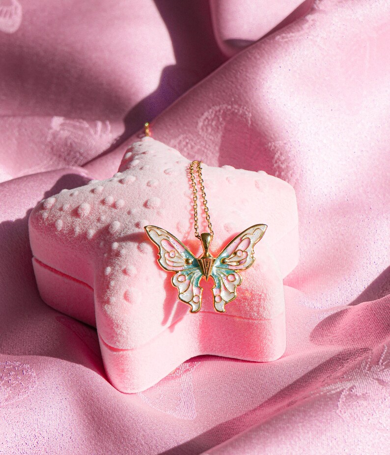 Pu&ograve; includere: Una collana con ciondolo a forma di farfalla dorata con dettagli in smalto blu e bianco, riposante in una scatola a forma di stella in velluto rosa.
