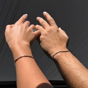 May include: Two people wearing simple silver beaded bracelets on their wrists. The bracelets are thin and delicate, with small silver beads spaced evenly along the length of the bracelet.