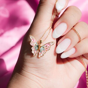Pu&ograve; includere: Una collana con catena dorata con un ciondolo a forma di farfalla rosa e blu. La farfalla ha un centro bianco e dettagli dorati. La collana &egrave; tenuta da una mano con smalto bianco.