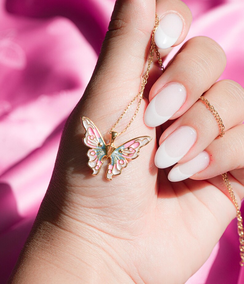 Pu&ograve; includere: Una collana con catena dorata con un ciondolo a forma di farfalla rosa e blu. La farfalla ha un centro bianco e dettagli dorati. La collana &egrave; tenuta da una mano con smalto bianco.
