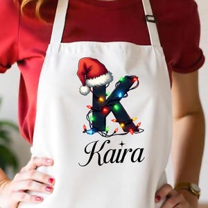 May include: White apron featuring a Christmas-themed design. The letter "K" is adorned with colorful Christmas lights and a Santa hat. The name "Kaira" is written in elegant script below the letter.