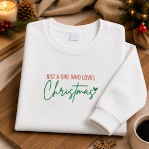 May include: A white crewneck sweatshirt featuring embroidered text in red and green. The text reads JUST A GIRL WHO LOVES in red block letters, followed by the word Christmas in a green cursive script with a small green heart icon at the end.