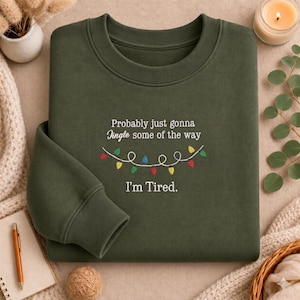 May include: An olive green crewneck sweatshirt featuring white embroidered text that reads "Probably just gonna Jingle some of the way" and "I'm Tired." Below the first line of text is an embroidered string of colourful Christmas lights.