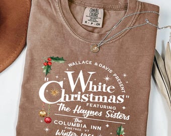 May include: A brown Comfort Colors t-shirt with the text "White Christmas" featuring The Haynes Sisters. The design includes holly and ornaments. A silver necklace with a pendant is draped across the shirt.