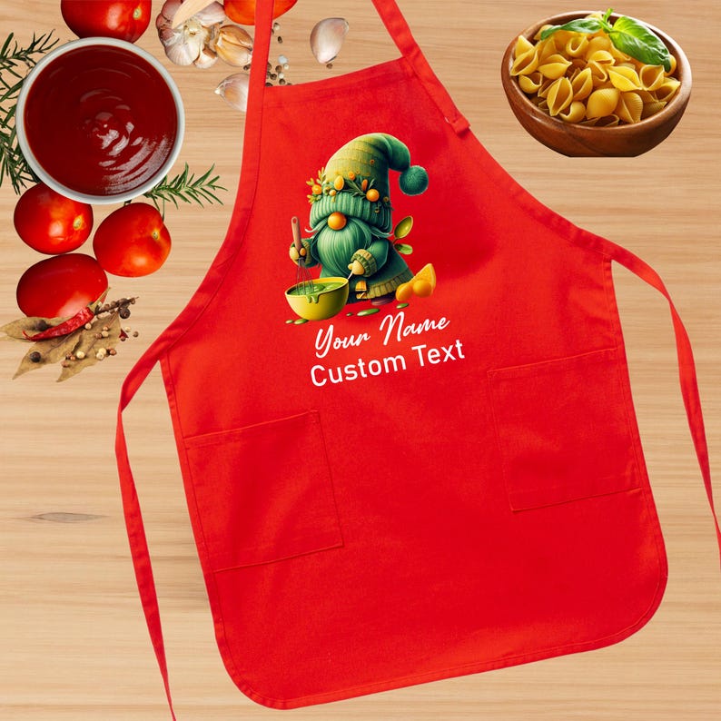 May include: Red kitchen apron with a green gnome holding a bowl and a wooden spoon. The apron has two pockets and the text "Your Name Custom Text" is printed on the front.