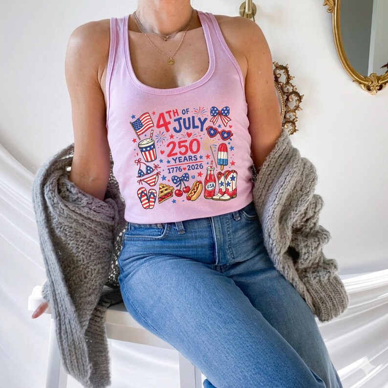 A woman wearing a pink tank top with a graphic design celebrating the 4th of July, along with a gray sweater, sits on a chair.