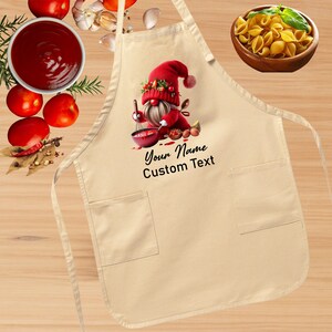 May include: A beige kitchen apron with a gnome wearing a red hat and holding a whisk. The gnome is surrounded by red sauce, eggs, and other ingredients. The apron has two pockets and the text "Your Name Custom Text" is printed on the front.