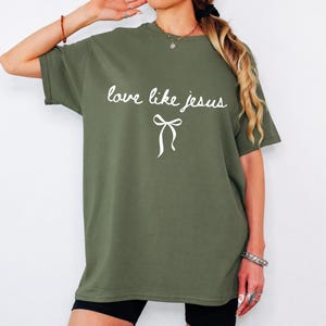 May include: An olive green short-sleeved t-shirt featuring the white text "love like jesus" in a cursive script font, positioned above a simple white line-drawn bow graphic. The shirt has a crew neckline and a relaxed, loose-fitting silhouette.