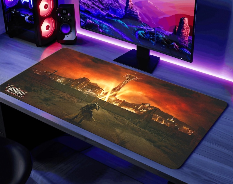 K&ouml;nnte beinhalten: Eine gro&szlig;e, rechteckige, braune und orangefarbene Gaming-Mausmatte mit einem Fallout 4-Logo und einer futuristischen Stadtlandschaft. Die Mausmatte liegt auf einem Schreibtisch mit einem Computermonitor und Lautsprechern.