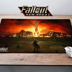 May include: A Fallout: New Vegas themed mouse mat with a red, orange and yellow sunset background. The image features a lone figure standing in the foreground, with the iconic New Vegas Strip in the background. The mouse mat has the Fallout: New Vegas logo in the bottom left corner.