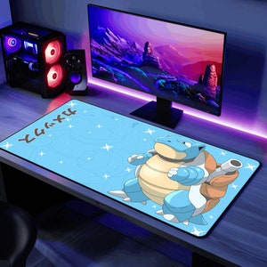 May include: A blue and brown cartoon character with a shell on its back, on a blue and white gaming mouse pad with the Japanese text "Squirtle" in black. The mouse pad is on a desk with a computer monitor and other electronics.