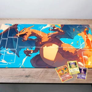 May include: A blue and orange Pokemon playing card game mat featuring a large illustration of Charizard, a fire-type Pokemon. The mat has a grid pattern for organizing cards. Three Pokemon trading cards are also shown on the mat.