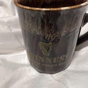 May include: A dark brown ceramic mug with a glossy drip glaze finish at the rim. The front features a gold Guinness harp logo and the text GUINNESS and beer comes near printed in gold. The mug has a curved handle on the right side.