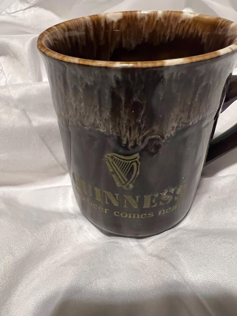 May include: A dark brown ceramic mug with a glossy drip glaze finish at the rim. The front features a gold Guinness harp logo and the text GUINNESS and beer comes near printed in gold. The mug has a curved handle on the right side.