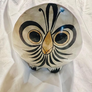 Op de afbeelding: Een afgerond keramisch beeldhouwwerk in de vorm van een uil met een crèmekleurige basis. Het heeft grote, donkere ronde ogen met beige accenten, een centrale beige snavel en zwarte, stralende veerdetails die naar buiten wijzen.