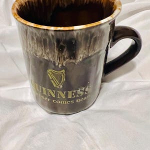 May include: A brown ceramic mug with a glossy drip glaze finish at the rim. The front features a gold harp logo and the text GUINNESS and no beer comes near. The mug has a curved handle on the right side and a smooth, dark brown body.