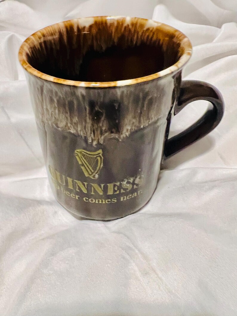 May include: A brown ceramic mug with a glossy drip glaze finish at the rim. The front features a gold harp logo and the text GUINNESS and no beer comes near. The mug has a curved handle on the right side and a smooth, dark brown body.