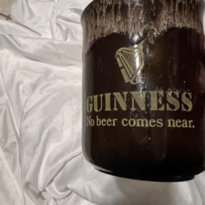 May include: A dark brown ceramic mug with a cream-colored drip glaze at the rim. The front features a gold-colored Guinness harp logo above the text "GUINNESS" and the phrase "No beer comes near." in gold lettering on the smooth, cylindrical body.