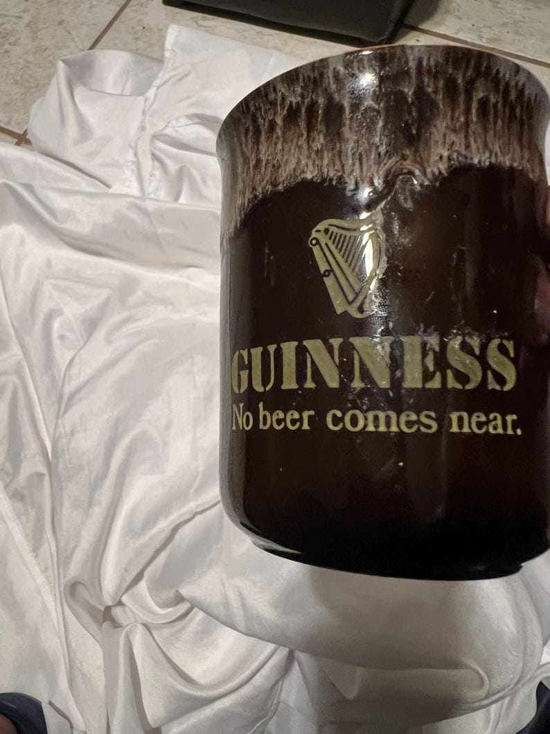 May include: A dark brown ceramic mug with a cream-colored drip glaze at the rim. The front features a gold-colored Guinness harp logo above the text "GUINNESS" and the phrase "No beer comes near." in gold lettering on the smooth, cylindrical body.