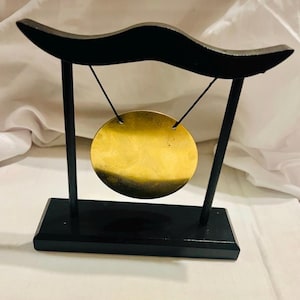 May include: A small, circular, metallic gold-colored gong suspended by two thin black cords from a black wooden frame. The frame consists of a rectangular base, two vertical supports, and a curved top bar. The gong has a smooth, reflective surface.