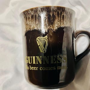 May include: A dark brown ceramic mug with a glossy finish and a cream-colored drip glaze at the rim. The mug features a gold harp logo, the text GUINNESS, and the phrase No beer comes near printed in gold lettering on the front side.