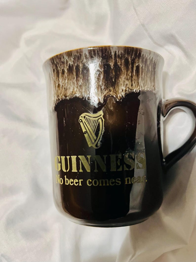 May include: A dark brown ceramic mug with a glossy finish and a cream-colored drip glaze at the rim. The mug features a gold harp logo, the text GUINNESS, and the phrase No beer comes near printed in gold lettering on the front side.