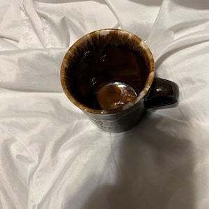 May include: A dark brown ceramic mug with a glossy finish and a distinctive drip-glaze effect around the rim, featuring a white and brown mottled pattern. The mug has a curved handle on the side and a deep, smooth interior, showcasing a classic design.