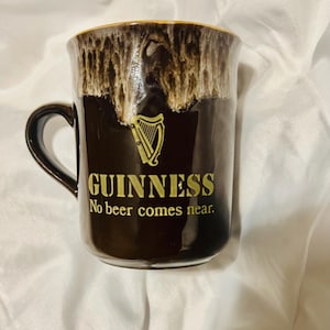 May include: A dark brown ceramic mug featuring a gold harp logo and the text "GUINNESS" followed by "No beer comes near." The upper rim has a decorative white and brown drip glaze effect, and the mug includes a curved handle on the left side.