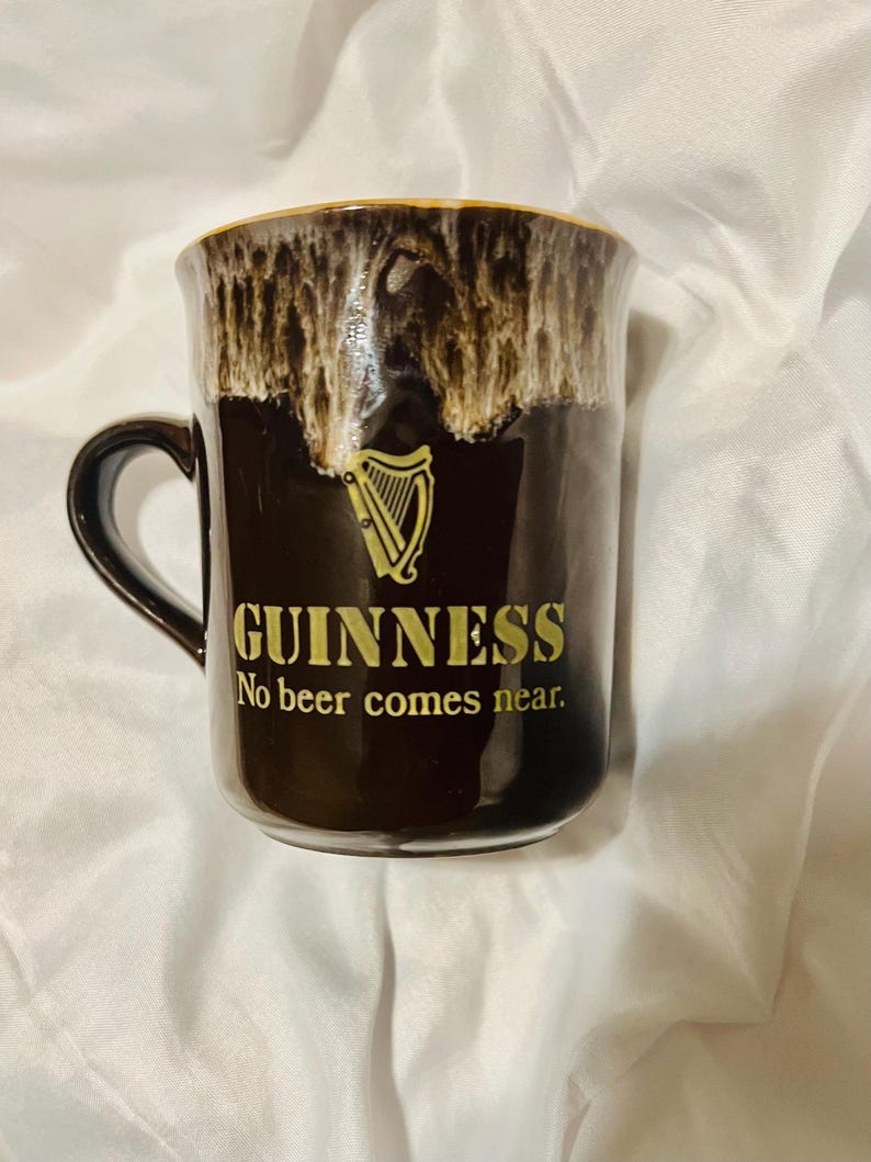 May include: A dark brown ceramic mug featuring a gold harp logo and the text "GUINNESS" followed by "No beer comes near." The upper rim has a decorative white and brown drip glaze effect, and the mug includes a curved handle on the left side.