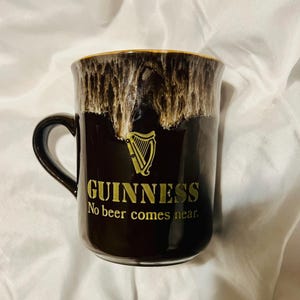 May include: A black ceramic mug featuring a gold harp logo and the text GUINNESS No beer comes near. The upper rim has a decorative drip glaze effect in shades of cream and brown, creating a textured appearance against the smooth black body.