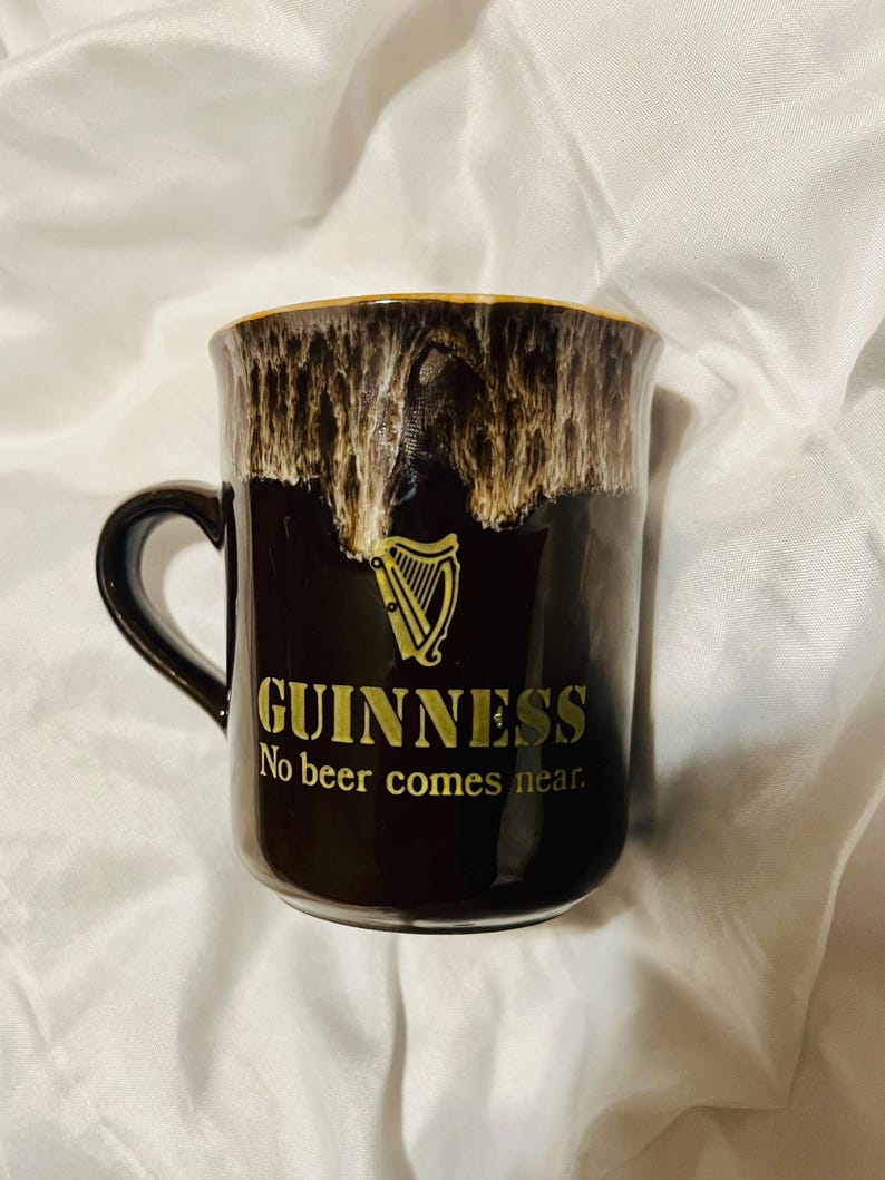 May include: A black ceramic mug featuring a gold harp logo and the text GUINNESS No beer comes near. The upper rim has a decorative drip glaze effect in shades of cream and brown, creating a textured appearance against the smooth black body.