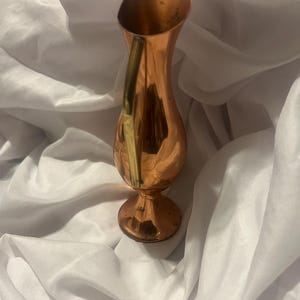 May include: A polished copper pitcher featuring a slender, curved neck, a bulbous body, and a flared circular base. The vessel includes a single vertical handle attached to the side, with a smooth, reflective metallic surface throughout the exterior.