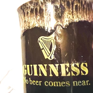 May include: A dark brown ceramic mug featuring a cream-colored drip glaze at the rim. The front displays a gold harp logo above the text GUINNESS and the slogan No beer comes near. The mug has a smooth, glossy finish and a cylindrical shape.