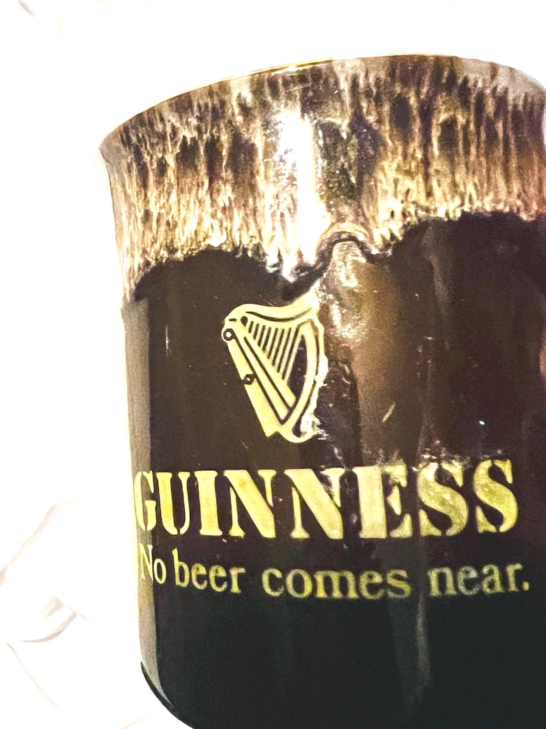 May include: A dark brown ceramic mug featuring a cream-colored drip glaze at the rim. The front displays a gold harp logo above the text GUINNESS and the slogan No beer comes near. The mug has a smooth, glossy finish and a cylindrical shape.