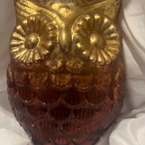 Puede incluir: Una figura decorativa de búho con un acabado dorado metálico en la parte superior y un tono cobre bruñido profundo en la parte inferior. El búho presenta grandes ojos circulares y un patrón de plumas festoneadas en toda su superficie.