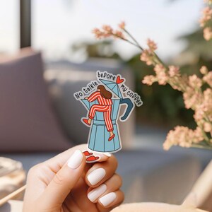 Puede incluir: Una pegatina con una ilustración de dibujos animados de una persona con pijama a rayas rojas y blancas subiendo a una cafetera azul. El texto de la pegatina dice: "No talkie before my coffee."
